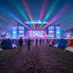 “Nighttime view of a lit EDM festival stage with neon light beams and silhouetted attendees walking through a camping area between installations.”